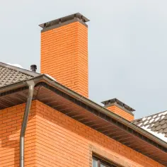 Ceglany komin i dach domu z rynnami, na tle jasnego nieba.